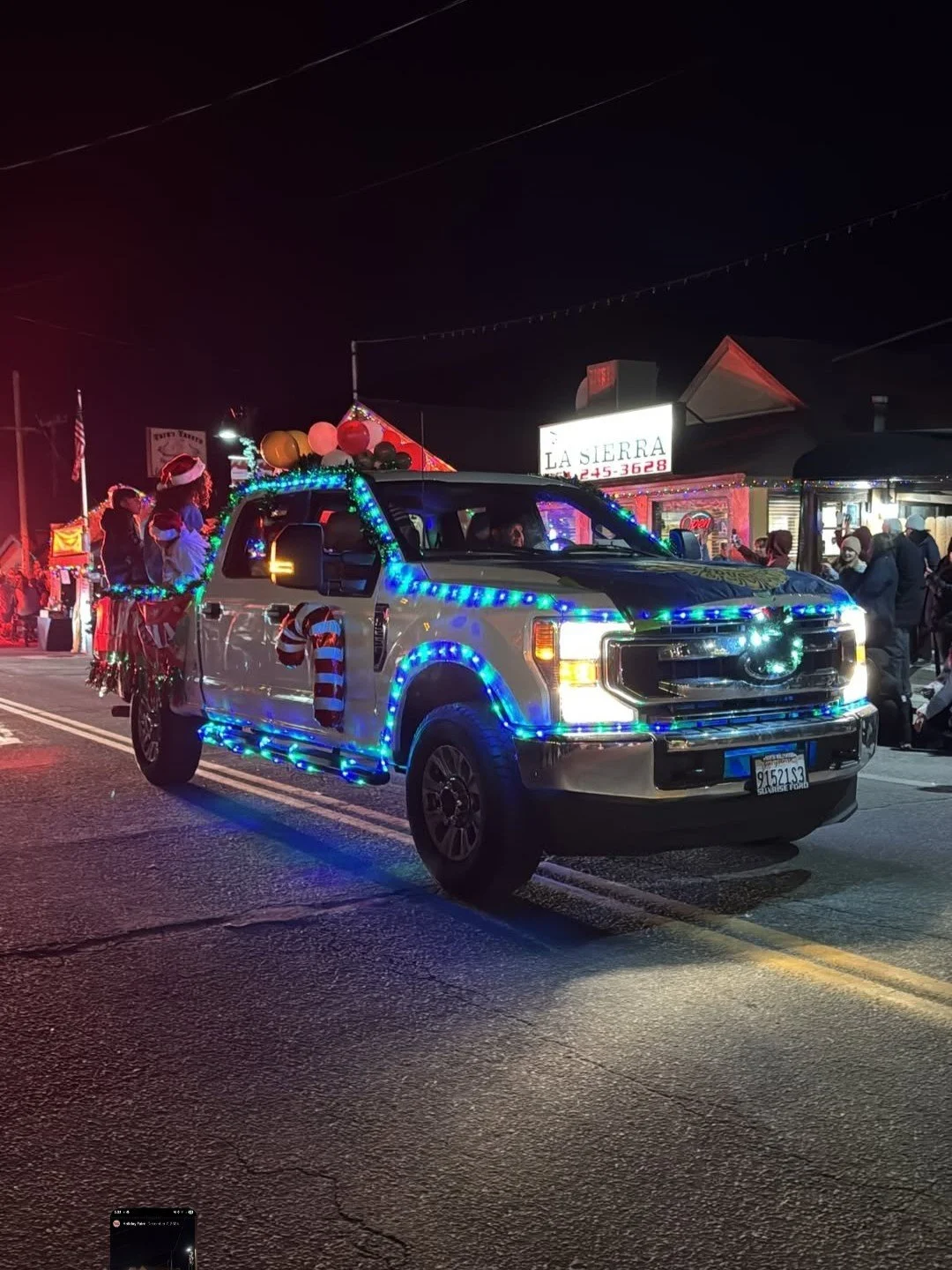 A pickup truck decorated with blue and white Christmas lights, with people dressed as Santa Claus and elves in the back, participating in a holiday parade on a city street at night.