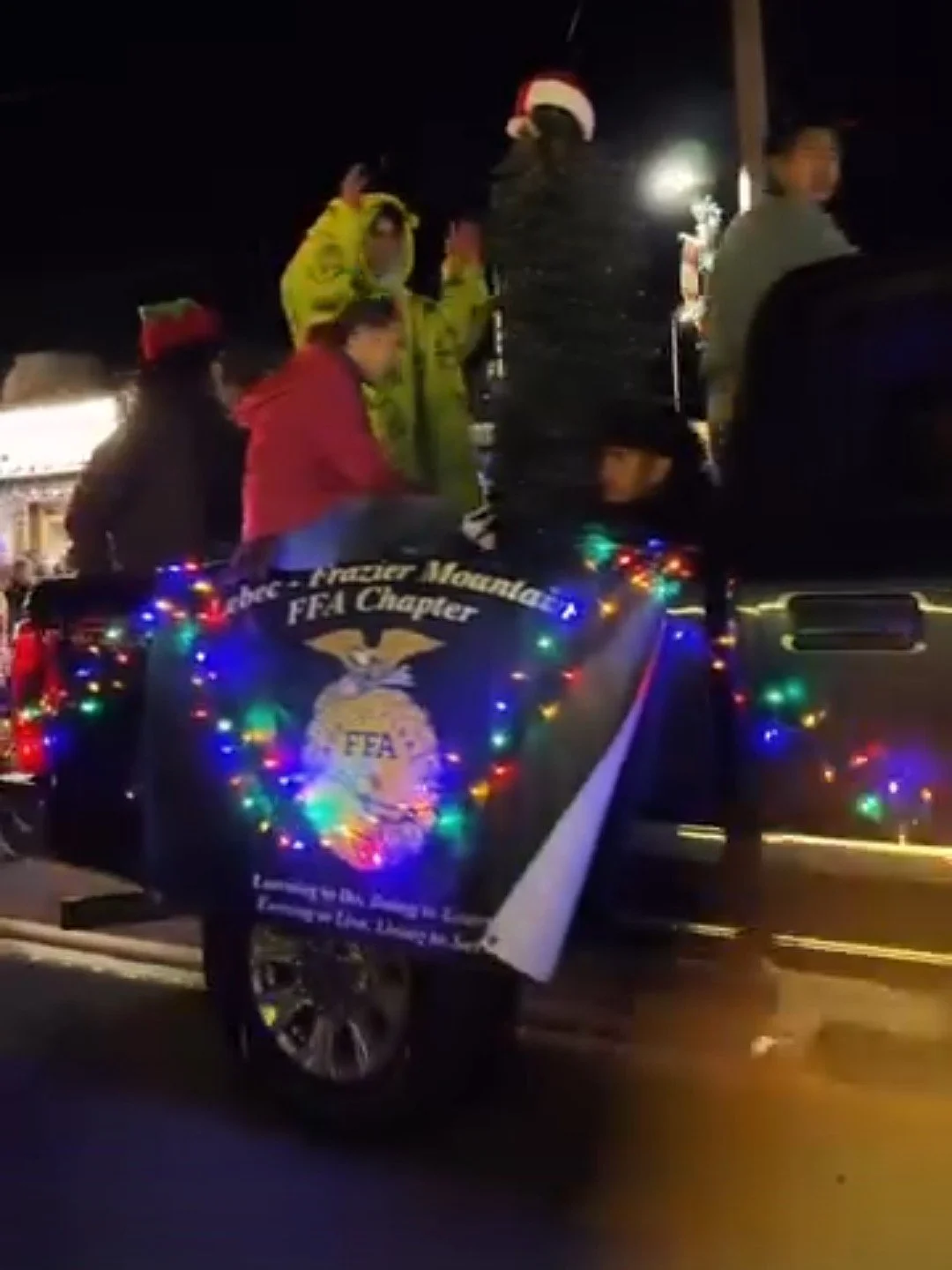 People riding in a decorated float with a sign for the FFA chapter, wearing winter clothing and Santa hats, during a nighttime parade.