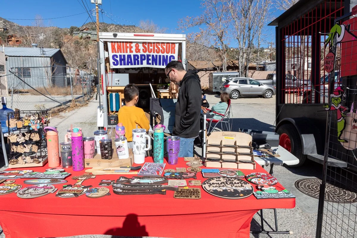Outdoor market stall selling knife and scissor sharpening services, with colorful mugs and various handcrafted items on display, and people browsing in an open area under clear blue skies.