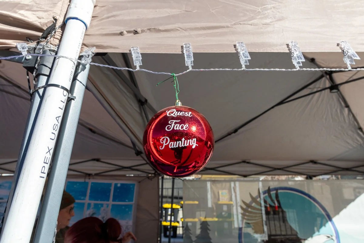 Red Christmas ornament decoration with the words "Quest Face Painting" hanging below a tent canopy at an outdoor event.