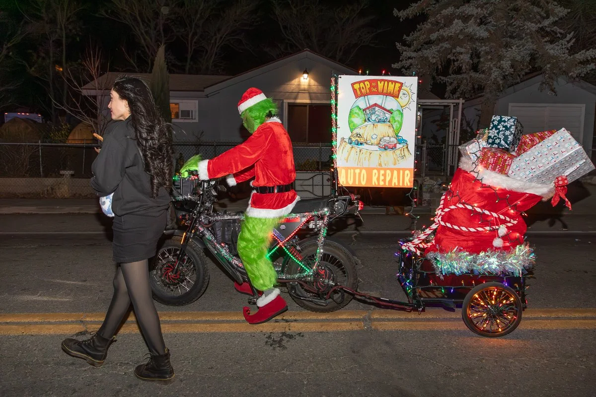 A person dressed as the Grinch riding a decorated electric bike pulling a cart filled with Christmas presents, while a woman walks in front of them on a nighttime street decorated for Christmas.