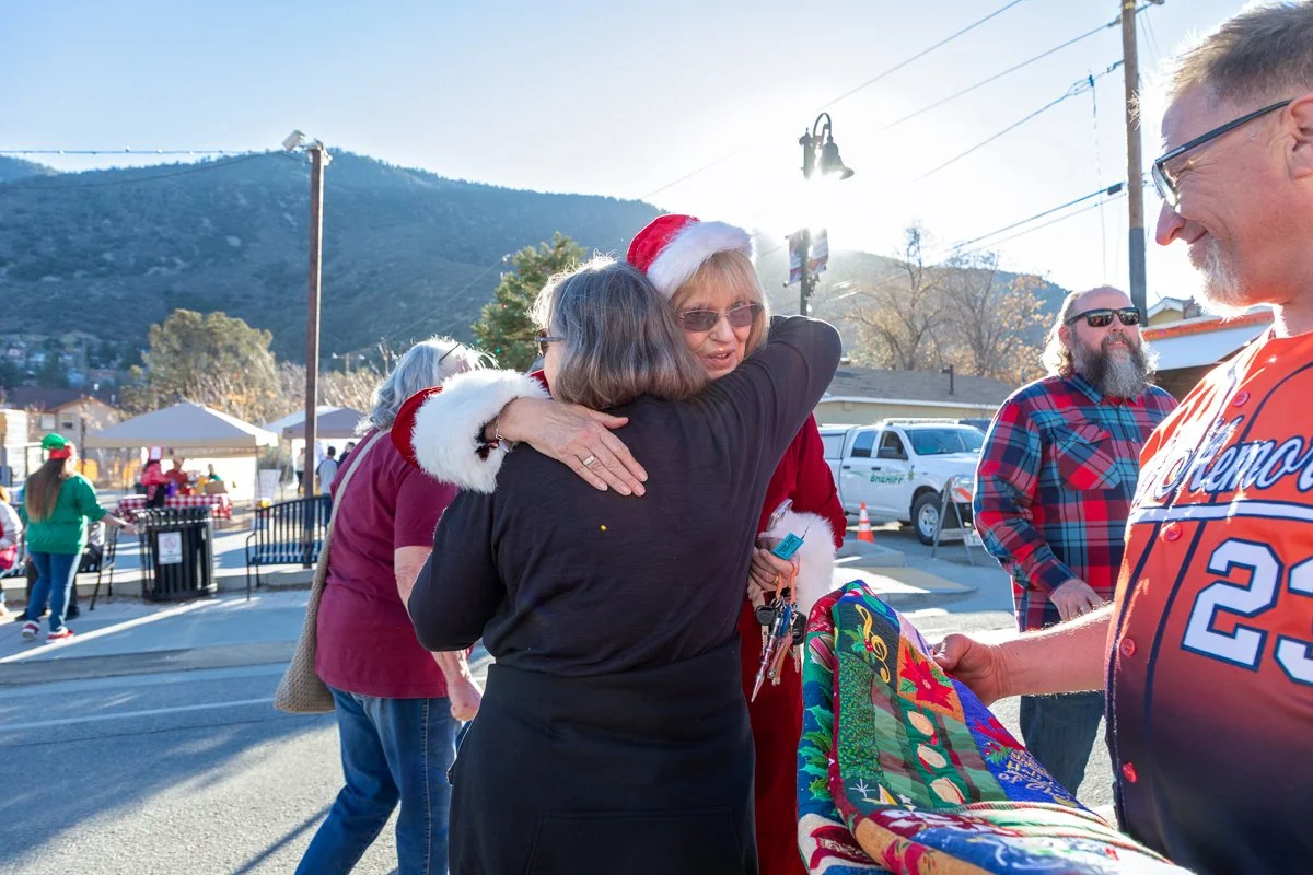 People celebrating Christmas outdoors, including a woman in a Santa hat hugging another woman, with others nearby exchanging gifts or socializing, and a mountain backdrop.