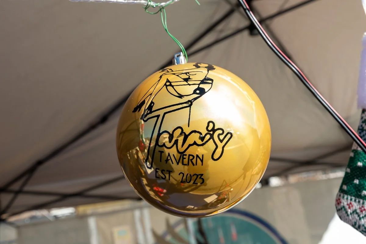 A gold Christmas ornament with black text and illustrations hanging under a tent. The text reads "Tara's Tavern EST 2023."