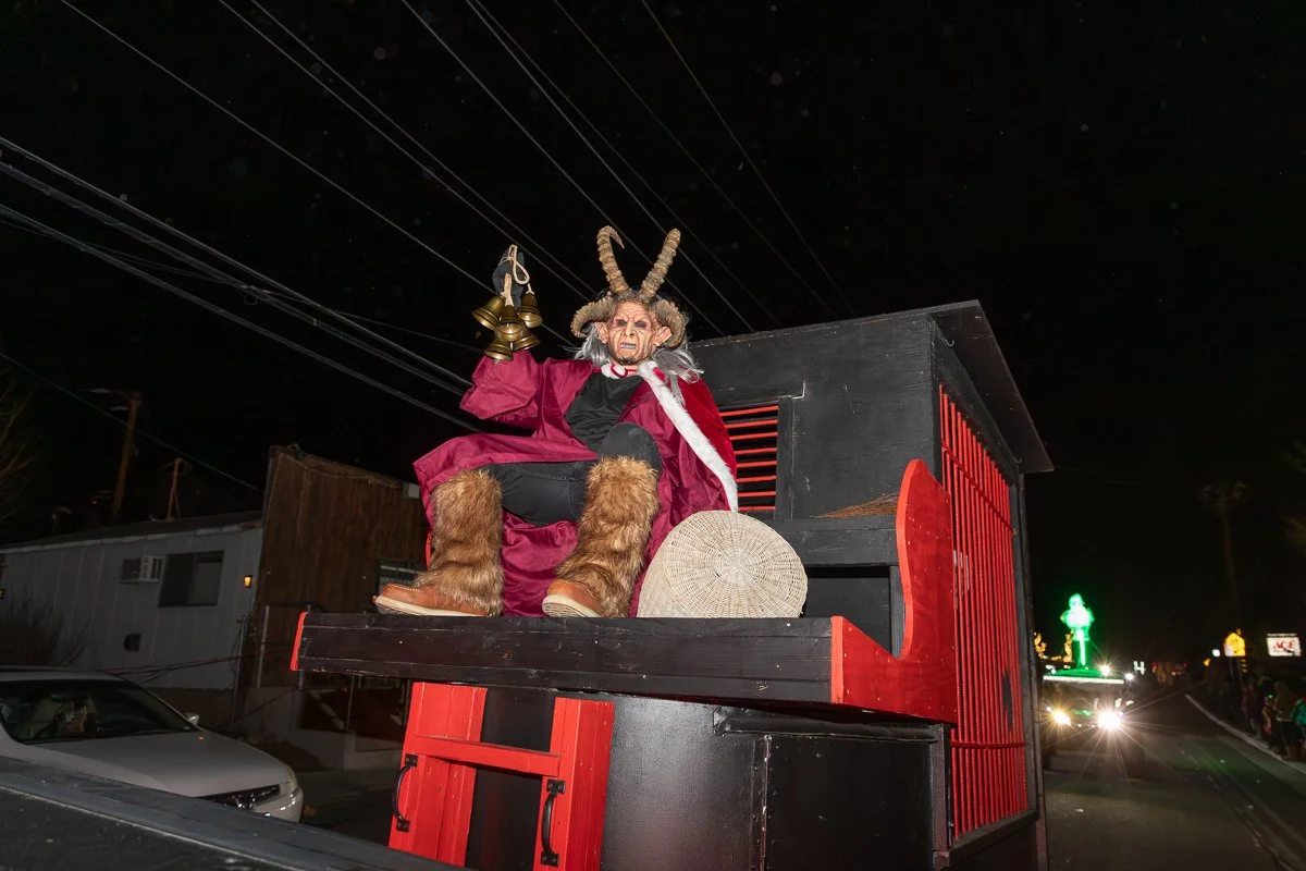 Decorated float featuring a figure of Krampus with horns, furry boots, and a red cloak, sitting on a black and red structure during a nighttime parade.
