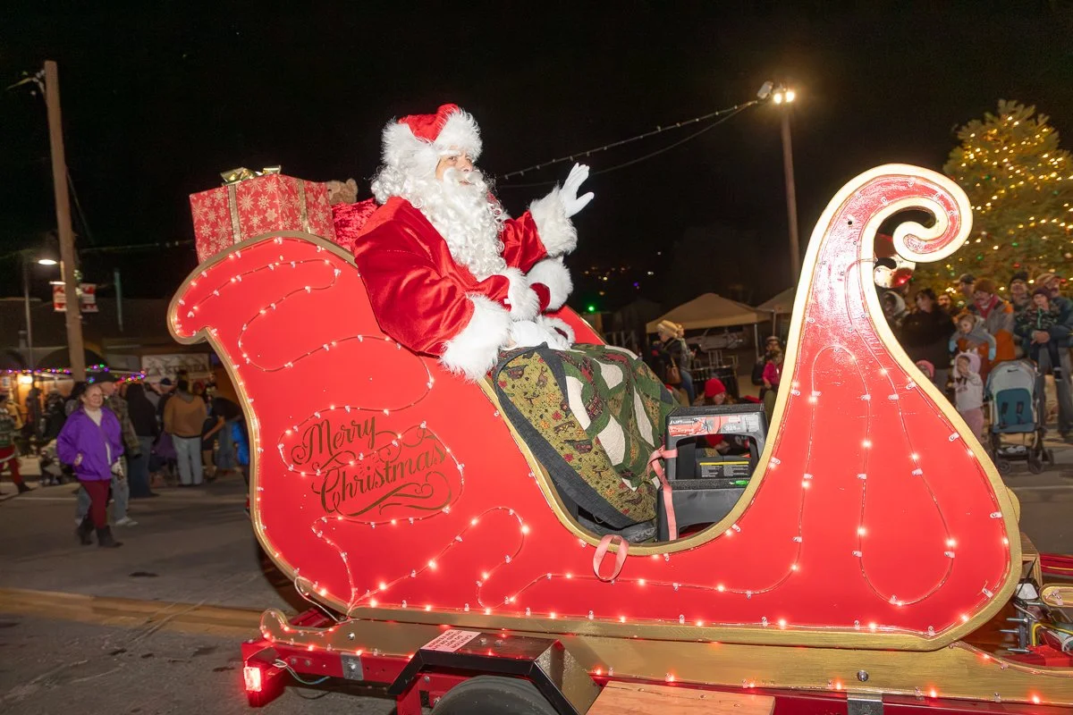 Santa Claus sitting in a decorated red sleigh with the words "Merry Christmas"; Santa waving at the crowd during a nighttime holiday parade, with people and Christmas lights in the background.