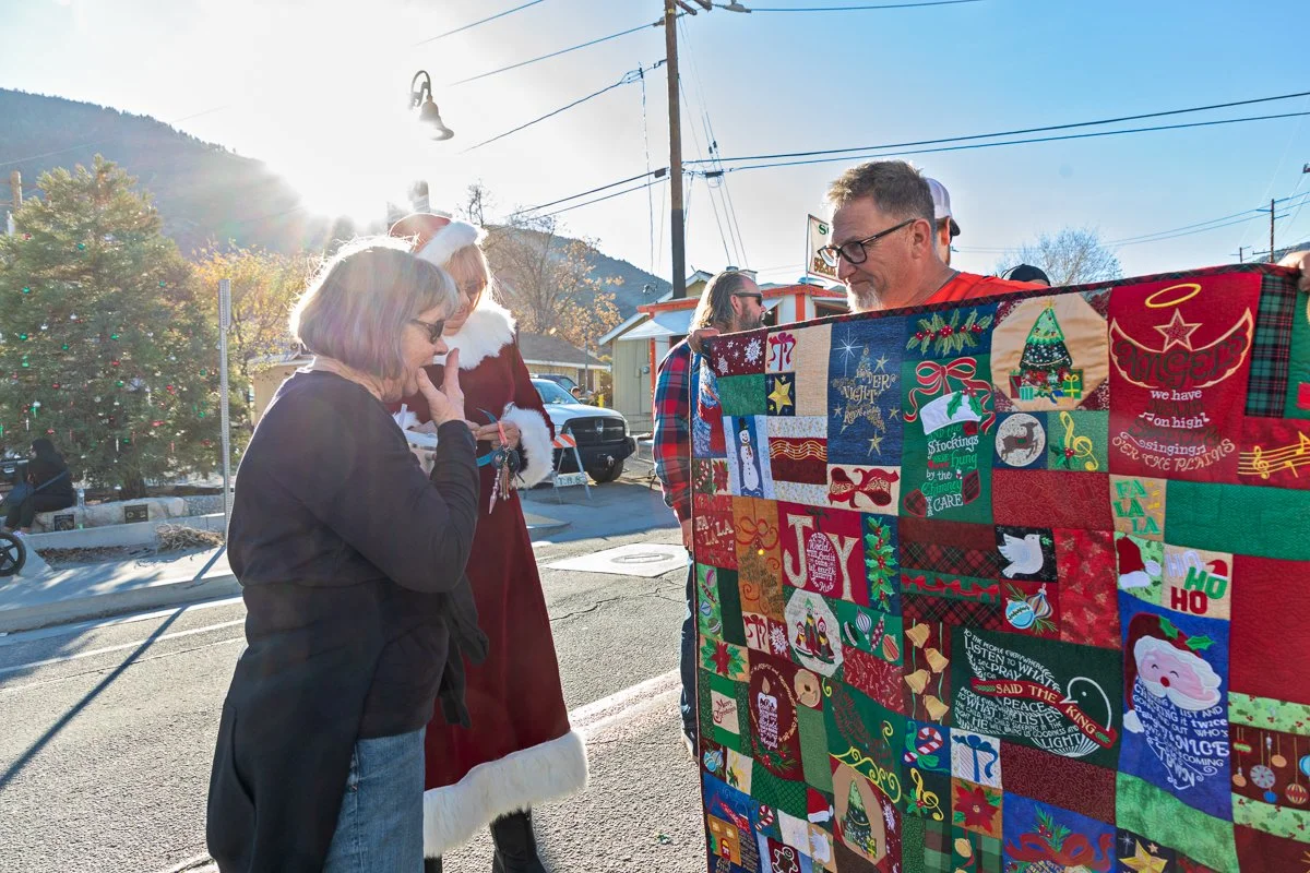 People looking at a large Christmas quilt outdoors during daytime. Two women, one in a Santa dress, and a man with glasses and a red plaid shirt hold the quilt. A Christmas tree decorated with ornaments is visible in the background.