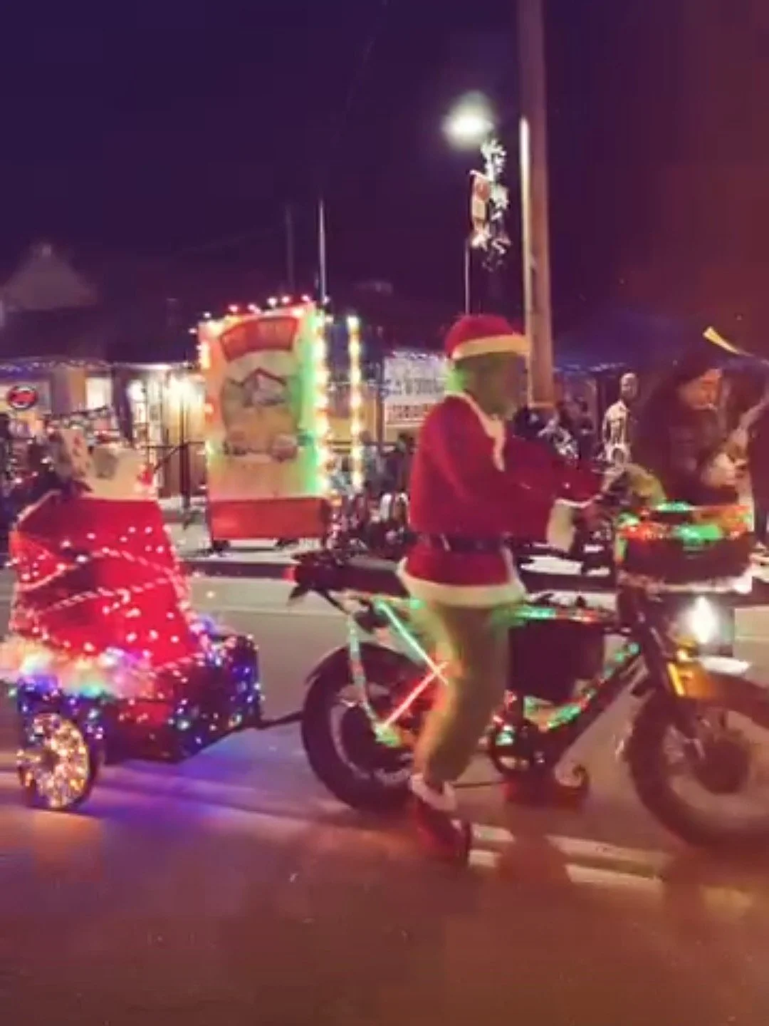 A night scene with people dressed in Christmas-themed costumes, including Santa suits and festive hats, riding bikes decorated with Christmas lights and ornaments. In the background, there are carnival rides and holiday decorations.