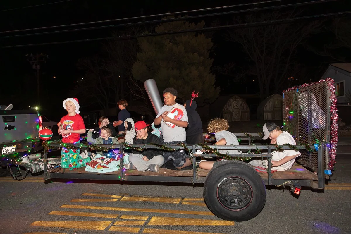 Children dressed in festive holiday costumes riding on a decorated flatbed trailer in a nighttime Christmas parade.