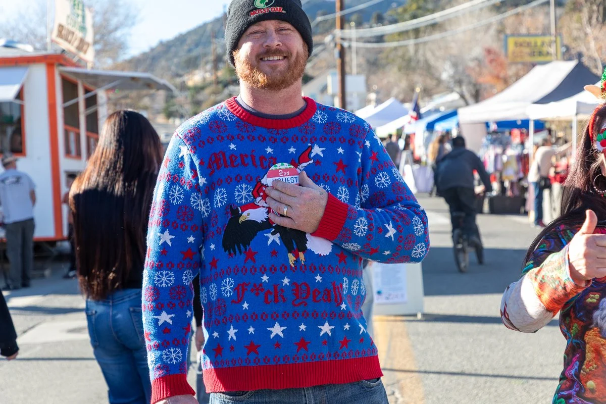 A smiling man in a festive sweater with an eagle and Christmas design, wearing a gray beanie, participating in an outdoor event or market.