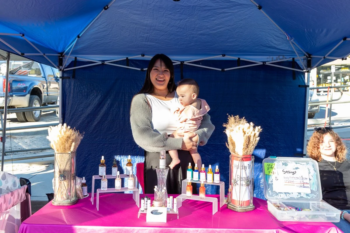 A woman smiling and holding a baby behind a booth at an outdoor market. The booth displays small bottles and decorative items on a pink tablecloth, with two vases of dried flowers on either side. A young girl with curly hair sits to the right in fron