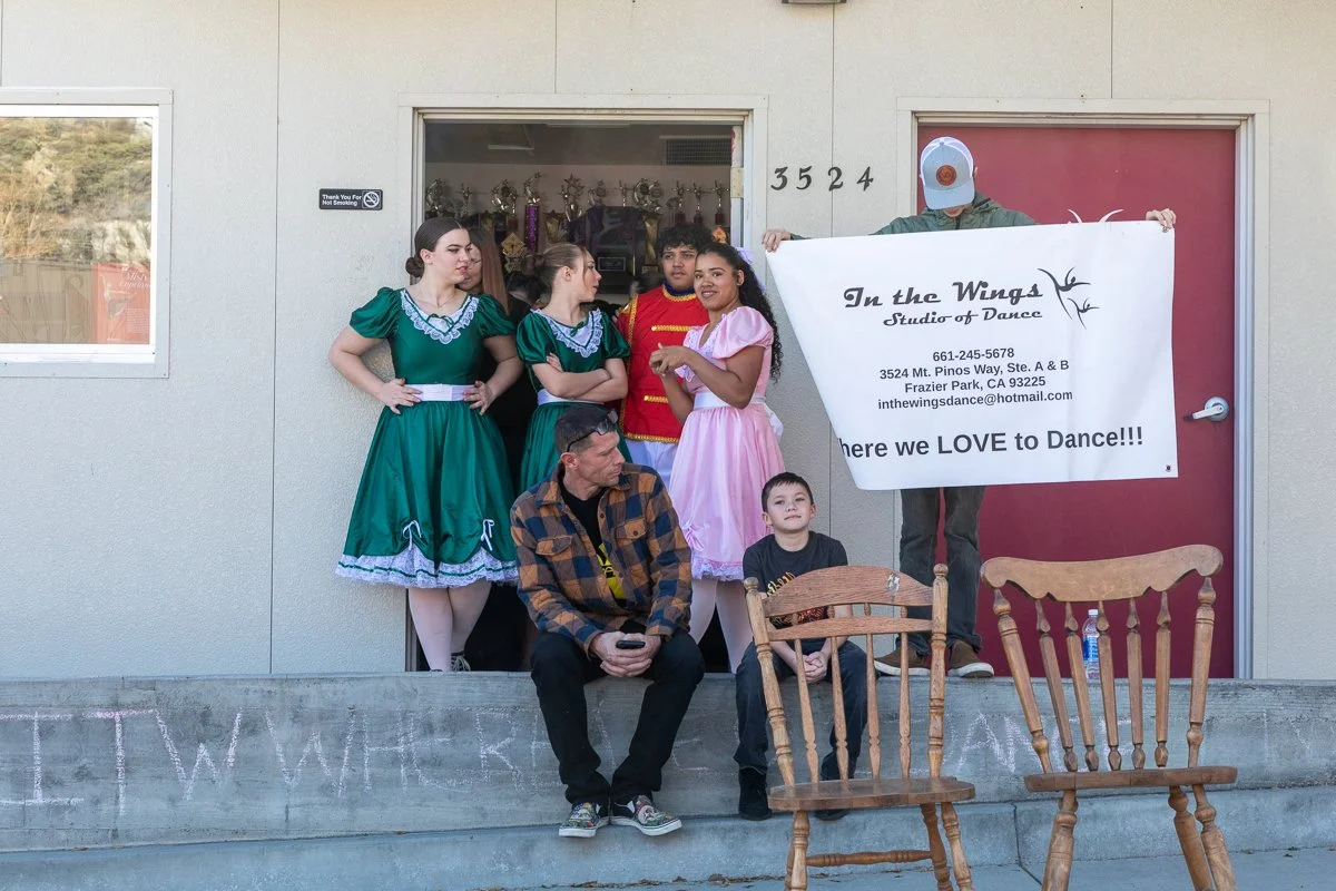 Group of people, some in colorful costumes, standing and sitting outside a dance studio with a sign that reads 'In the Wings Studio of Dance.' A person holds a large banner with studio information and a message about loving to dance. Two wooden chair