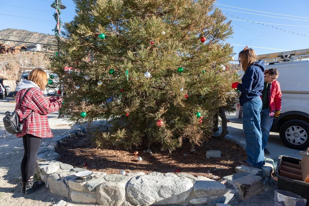 Three people decorate a large outdoor Christmas tree with ornaments, standing on rocks around the tree in winter clothing.