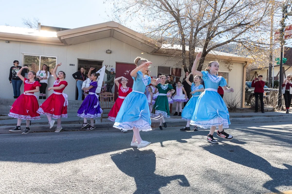 Children dressed in colorful vintage dresses performing a dance on a street during a parade or festival.