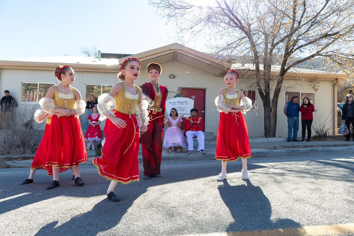 Group of young girls and a woman dressed in Russian-style costumes performing a dance on a street in front of an audience during daytime, with some onlookers and a building in the background.