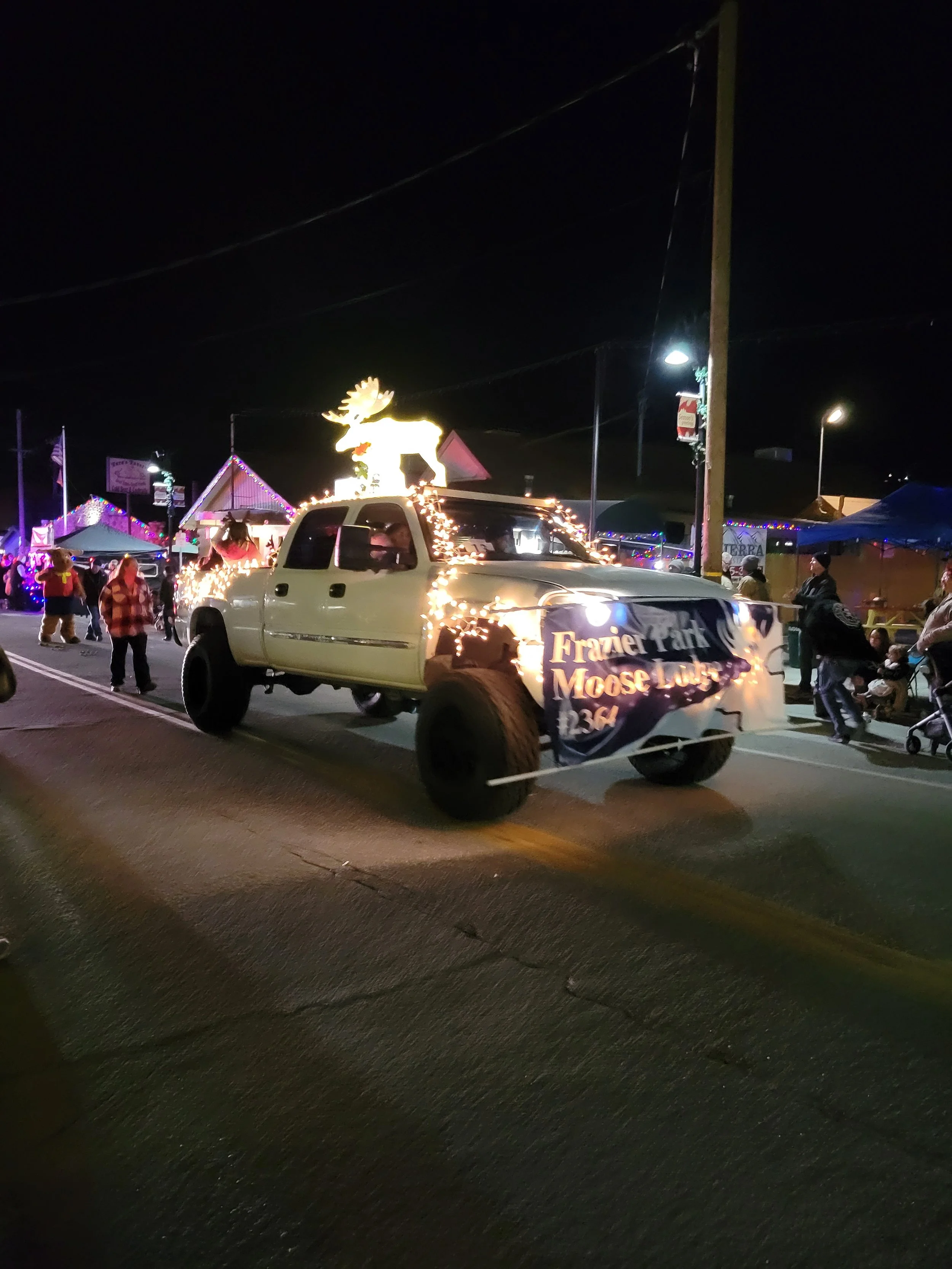 A decorated pickup truck in a nighttime parade with a moose figure on top, lights, and a banner reading "Frazier Park Moose Lodge 1364". There are people walking along the street and tents or stalls in the background.