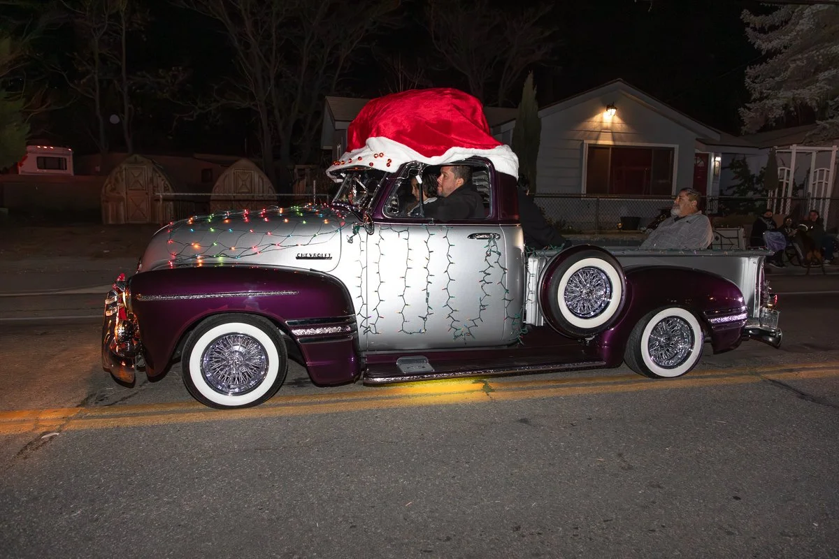 A vintage Chevrolet pickup truck decorated for Christmas with a Santa hat draped over the cab, Christmas lights on the hood, and a few people sitting inside and in the back of the truck during a nighttime parade.
