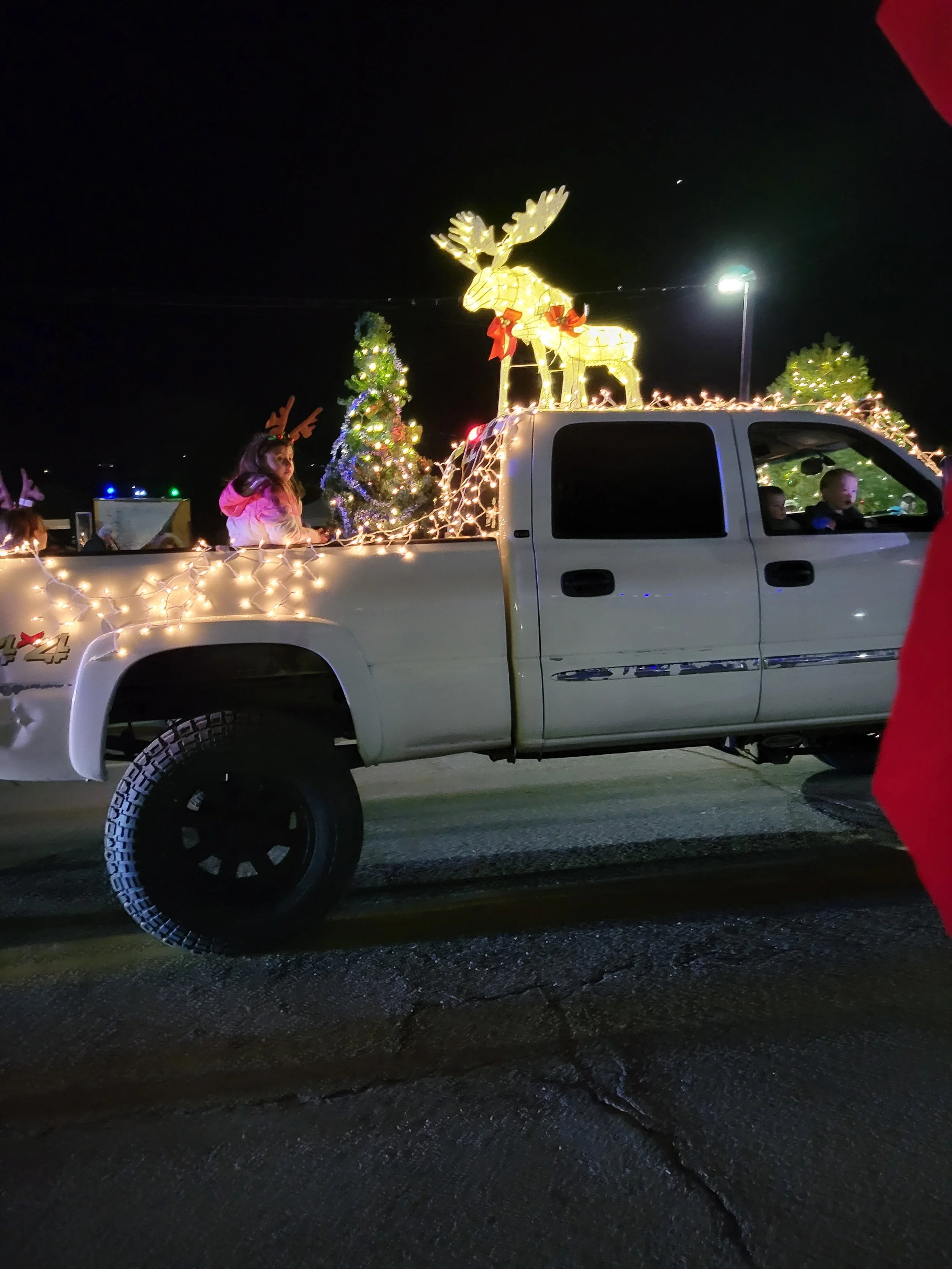 A white pickup truck decorated with Christmas lights, a small Christmas tree, and a large illuminated reindeer on top. Kids with reindeer antlers and winter clothing are in the back of the truck, celebrating during nighttime.