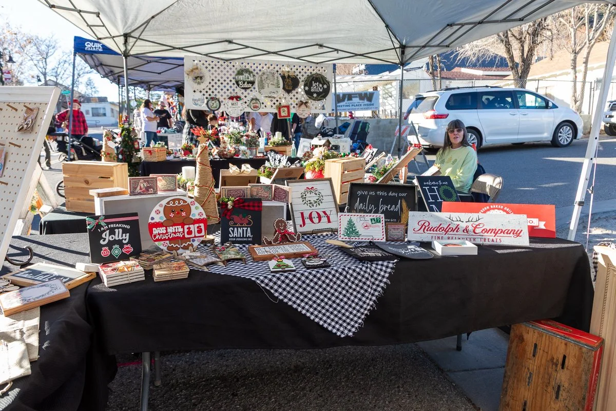 Outdoor holiday craft fair booth displays various Christmas-themed signs, decorations, and handmade items under a canopy, with a woman sitting behind the table and cars parked in the background.