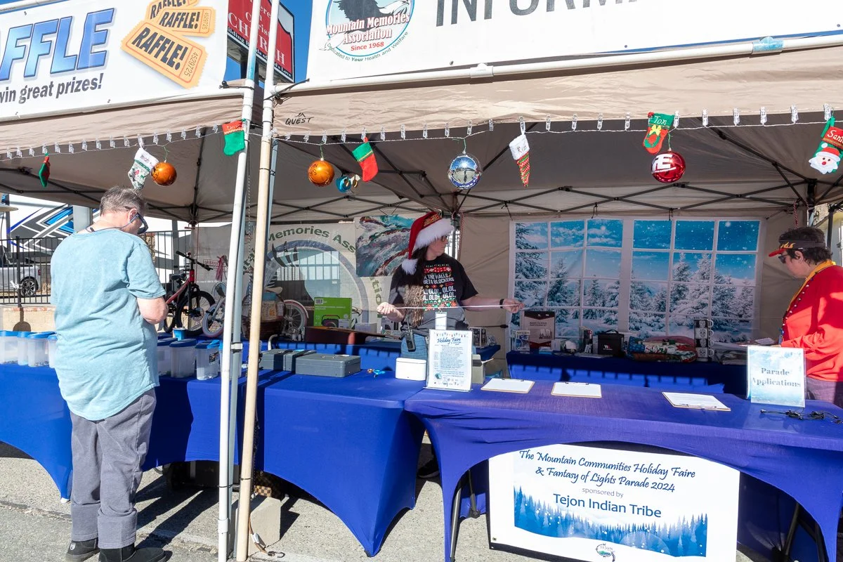 A booth at a Christmas event with decorations including ornaments, Christmas stockings, and a Santa hat on one person. People are standing at tables covered with blue tablecloths, and there is a backdrop of a snowy winter scene. The booth has signs f