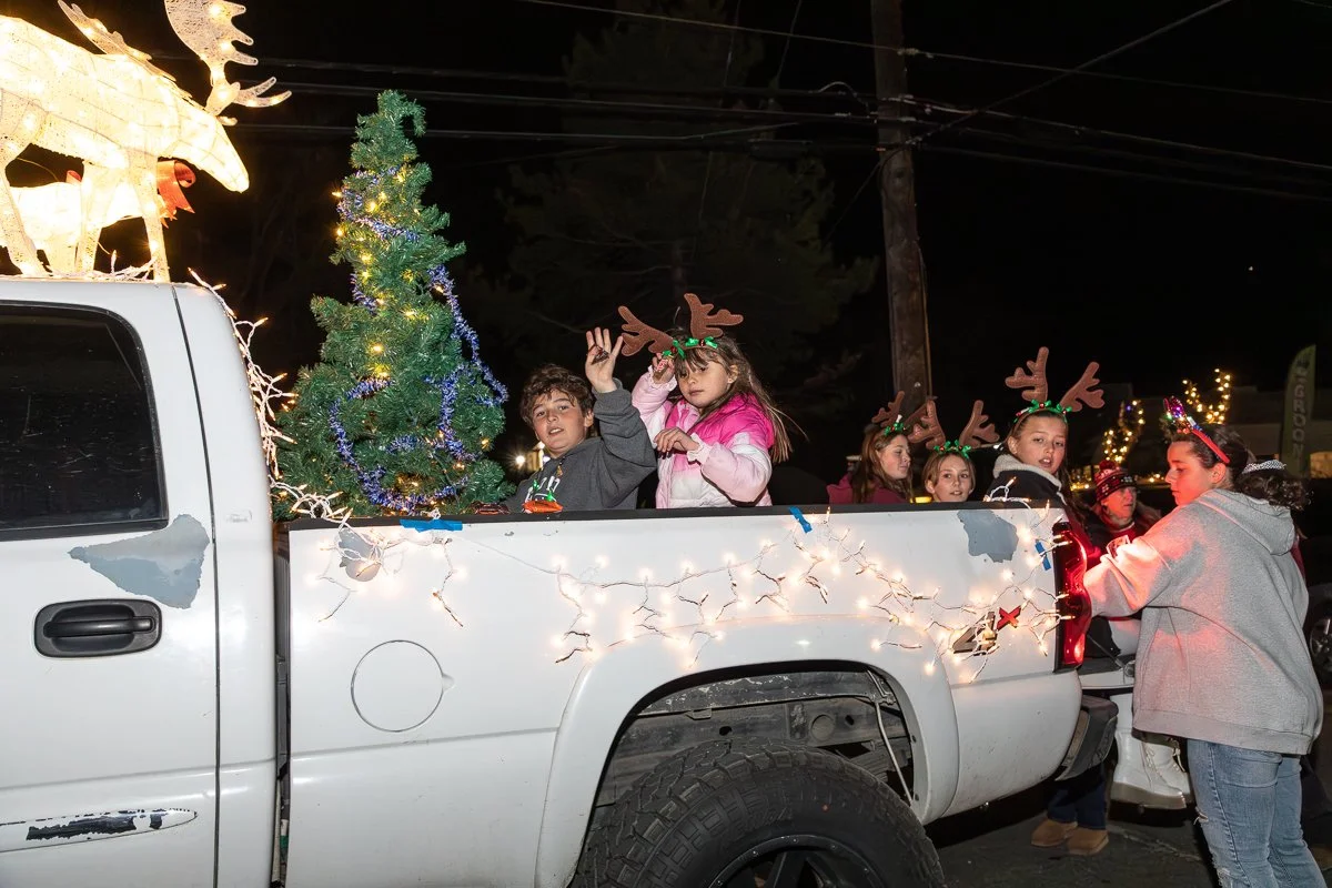 Children wearing reindeer antler headbands riding in the back of a decorated pickup truck during a nighttime Christmas parade. The truck has Christmas lights and a Christmas tree in the bed.
