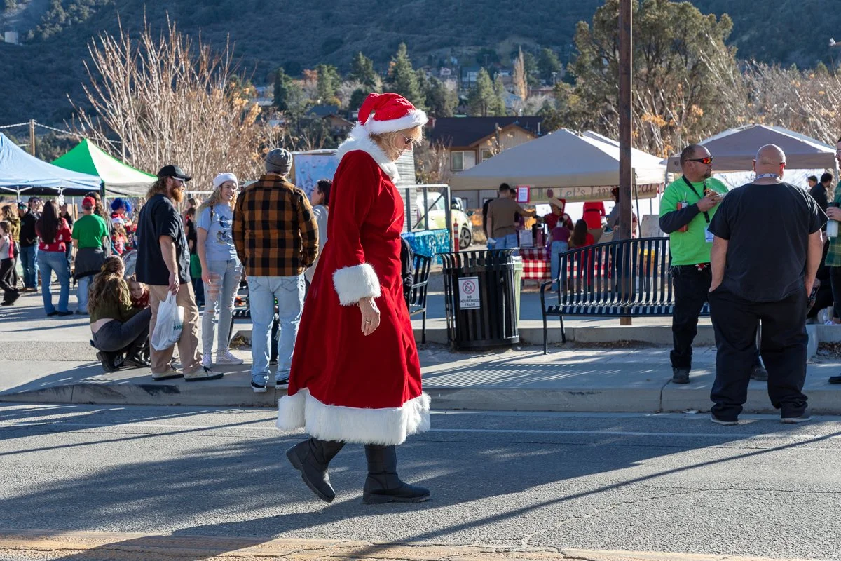 A woman dressed as Mrs. Claus walking at an outdoor holiday market with tents and people in the background, during the daytime.