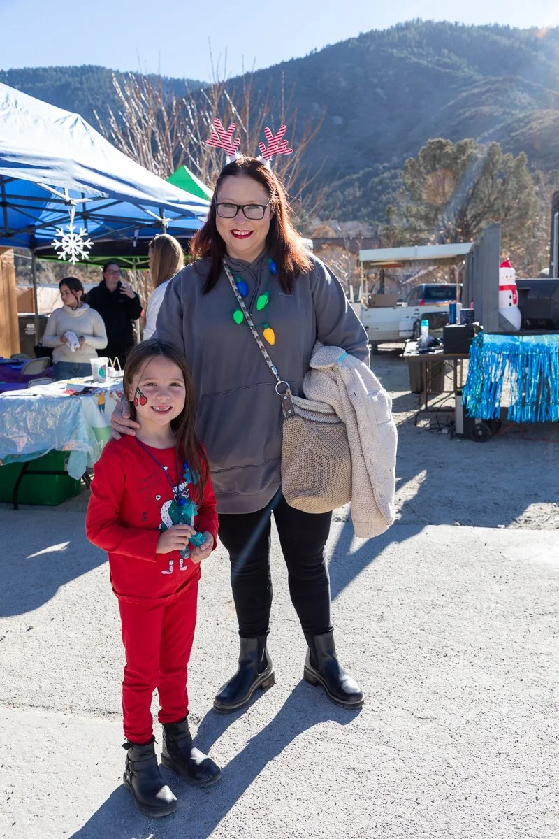 A woman and a young girl smiling outdoors at a holiday event. The woman is wearing reindeer antlers, glasses, a gray hoodie, and black boots, while the girl is dressed in a red festive outfit with holiday-themed face paint. In the background, there a