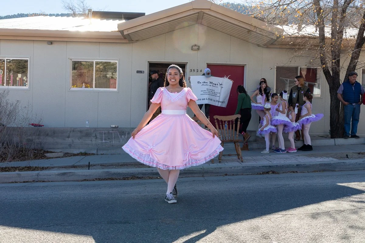 Young girl in a pink dress holding out the sides of her skirt, smiling, while a group of young girls in ballet costumes gather near a building in the background.