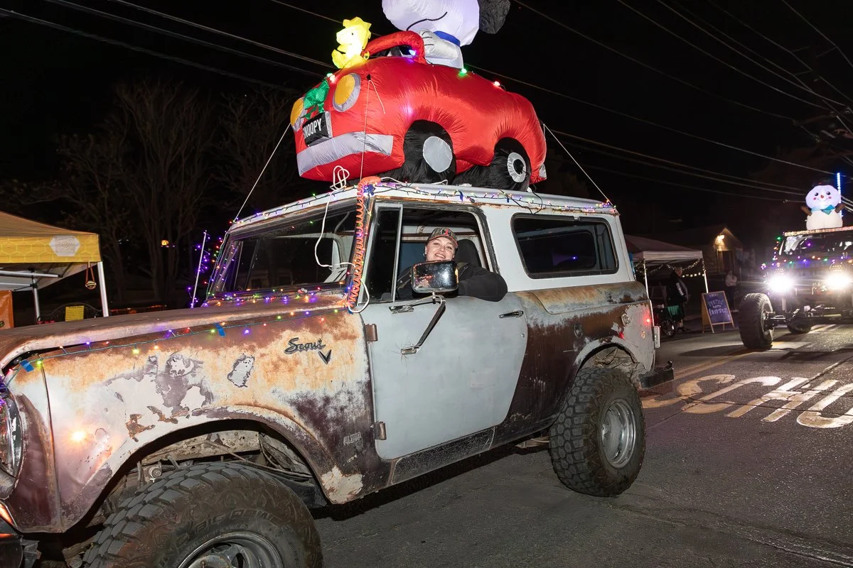 A vintage off-road vehicle decorated with Christmas lights, with a large inflatable Snoopy and his dog house on the roof, and a person smiling through the driver's side window at night.