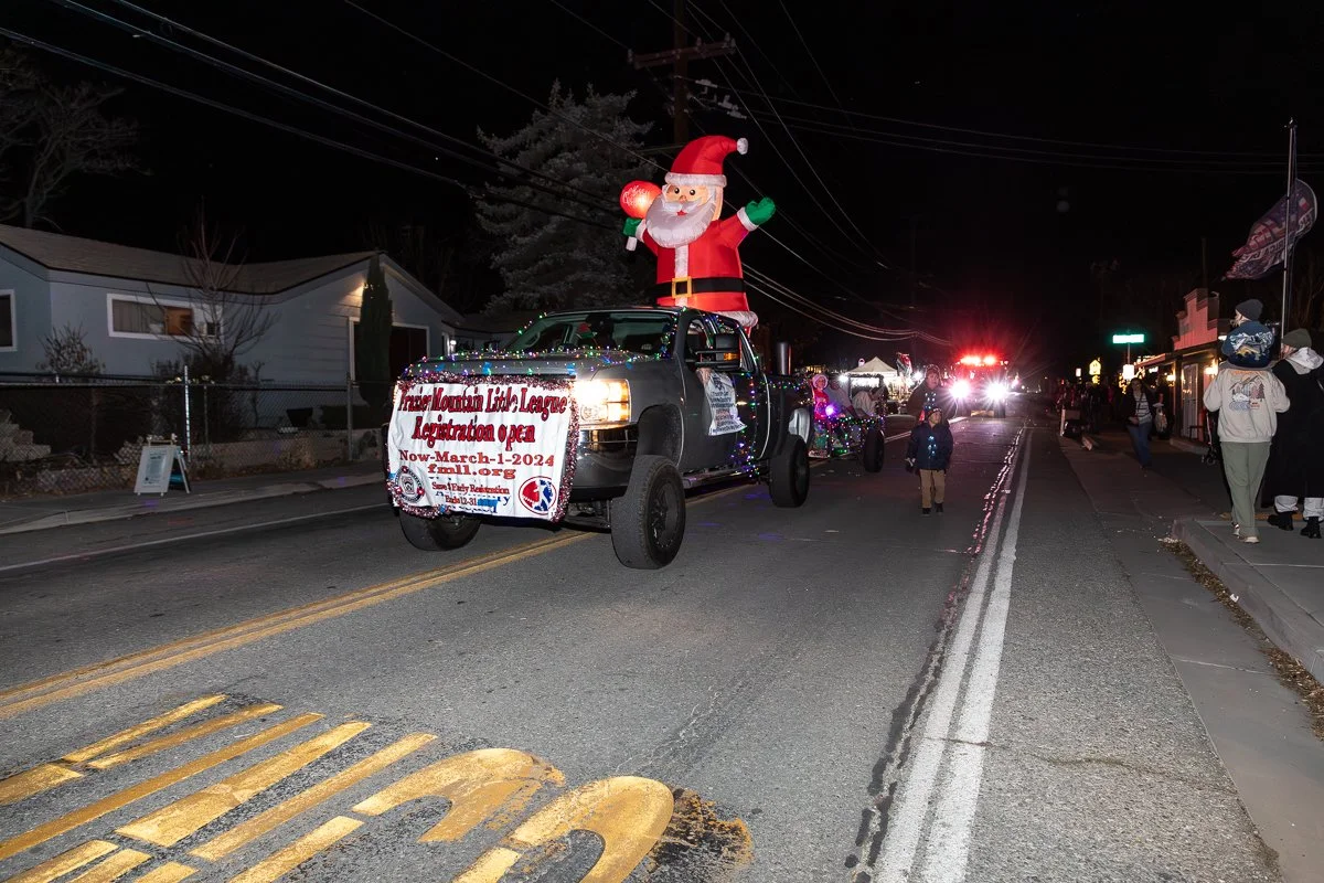 A nighttime Christmas parade on a street with a decorated float featuring an inflatable Santa Claus on top, surrounded by Christmas lights and a banner indicating the Transverse Mountain Lake League Lighting open event.