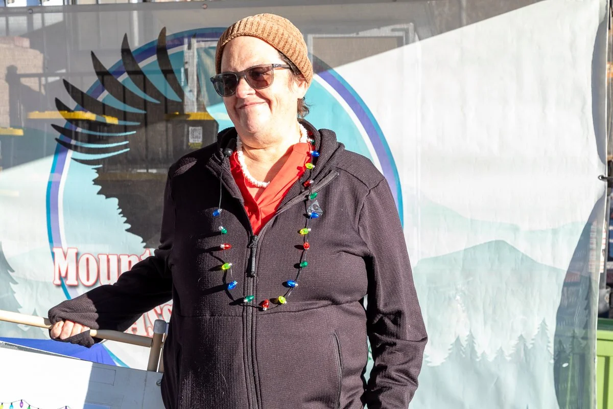 A woman wearing sunglasses, a brown knit beanie, a black jacket, an orange shirt, and a colorful bead necklace, standing outdoors in front of a banner with a mountain and wing design.