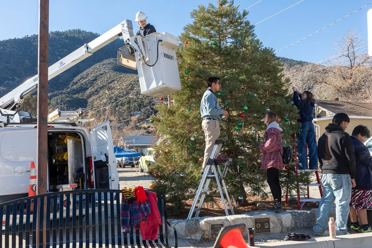 People decorating a large Christmas tree outdoors, with a cherry picker lift and utility truck in the background and mountains in the distance.