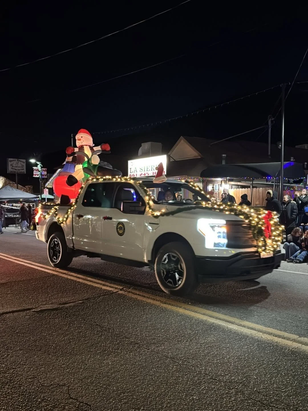 A white pickup truck decorated with Christmas lights and an inflatable Santa Claus on the bed, driving in a nighttime parade with people sitting and standing along the roadside.