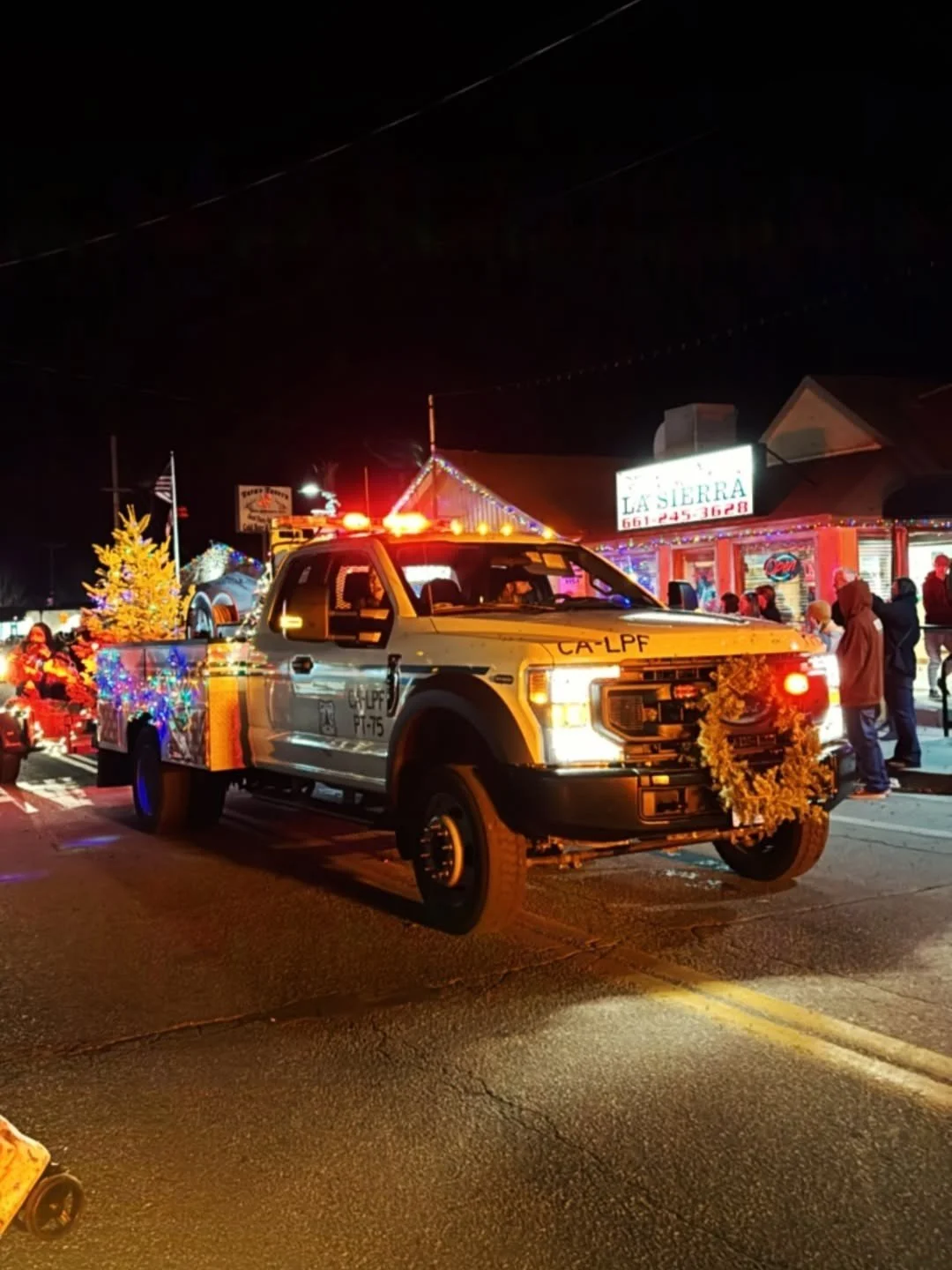 Decorated police vehicle with holiday garlands and lights, parked on a street at night during a festive event, with people gathered nearby and storefronts in the background.
