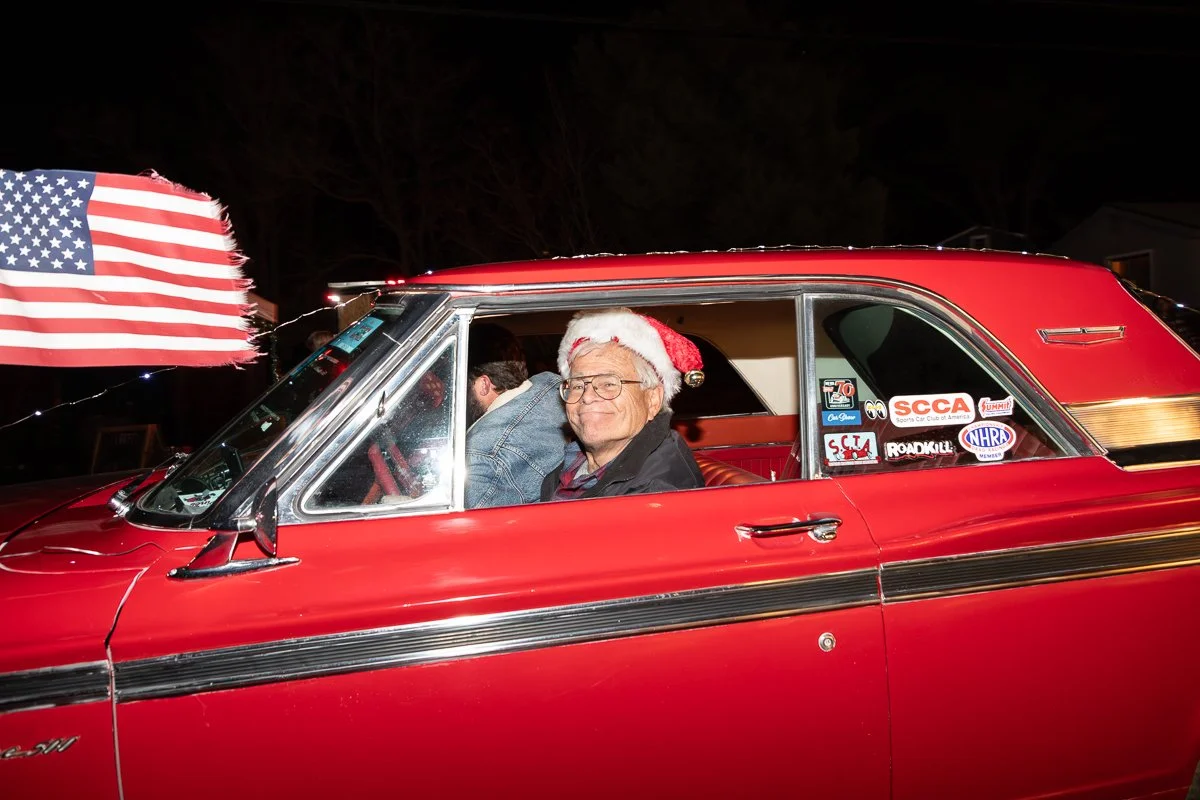 An elderly man with glasses and a Santa hat sitting in the driver's seat of a red vintage car, smiling. The car has a large American flag attached to the front left corner and is decorated with various automotive stickers on the side window. It is ni