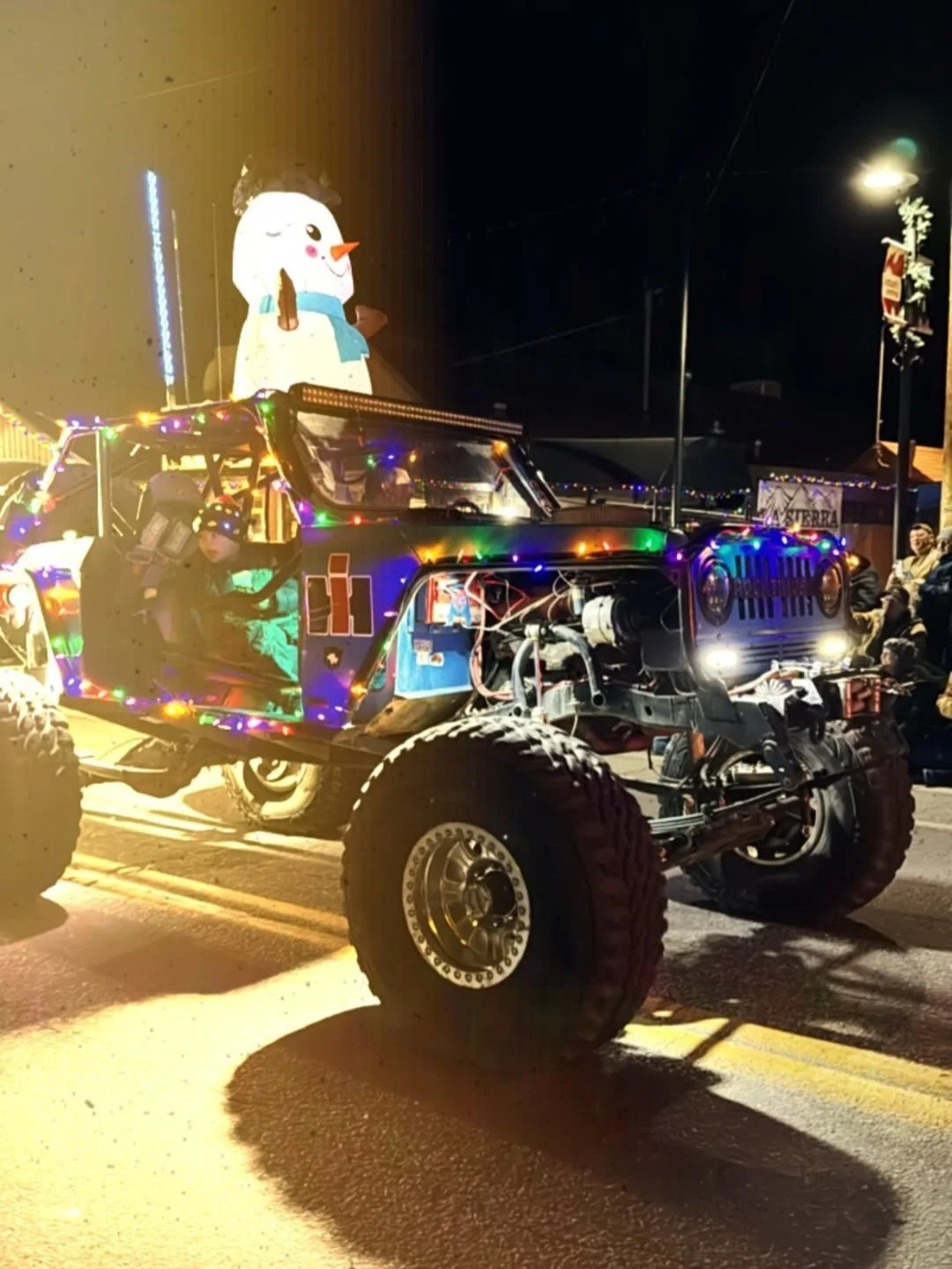 A decorated off-road vehicle (Jeep) with Christmas lights and a large snowman decoration on top, parked on a street at night during a parade or celebration, with people in the background.
