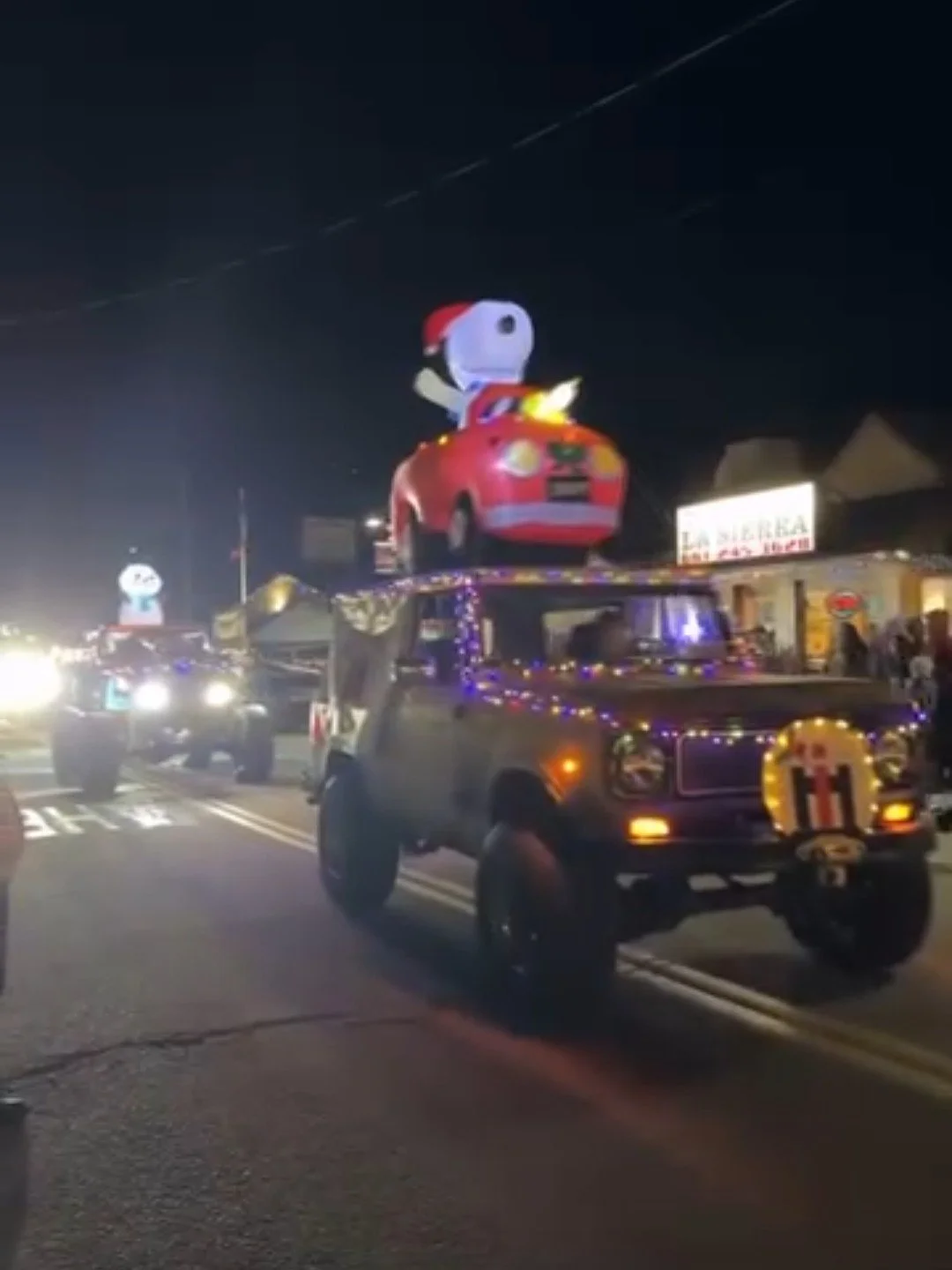 A decorated truck featuring a large Snoopy figure wearing a Santa hat and riding a red convertible, with colorful string lights, driving down a street at night during a parade or celebration.
