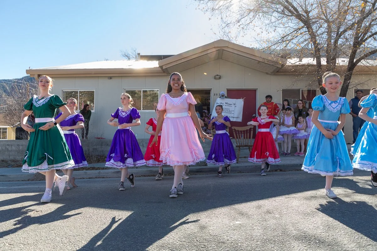 Young girls in colorful dresses performing a dance outdoors in front of a building with onlookers in the background.
