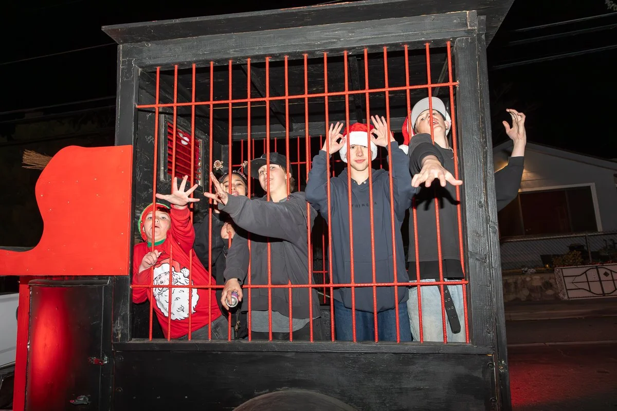 Group of people wearing Santa hats inside a red and black cage, reaching out through the bars, during a holiday event at night.