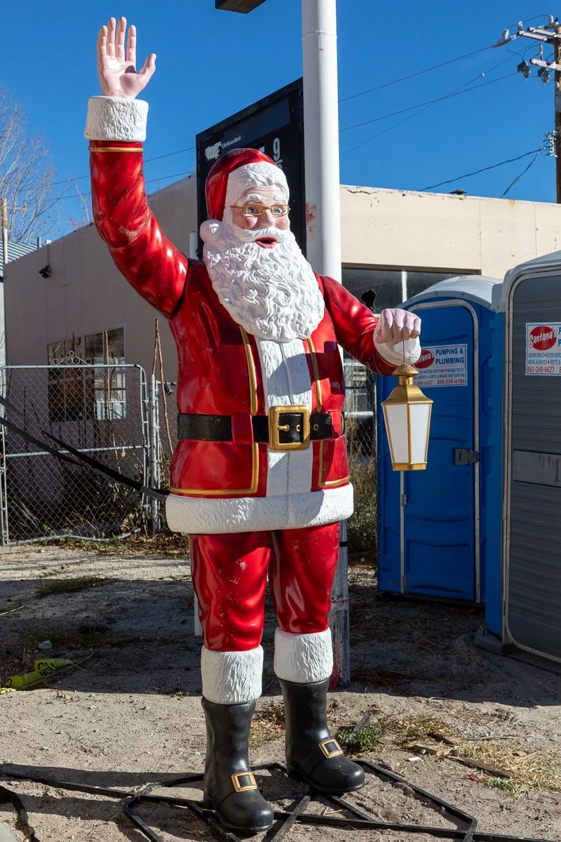Santa Claus statue waving, holding a lantern, outdoors with a clear blue sky, buildings, and portable toilets in the background.