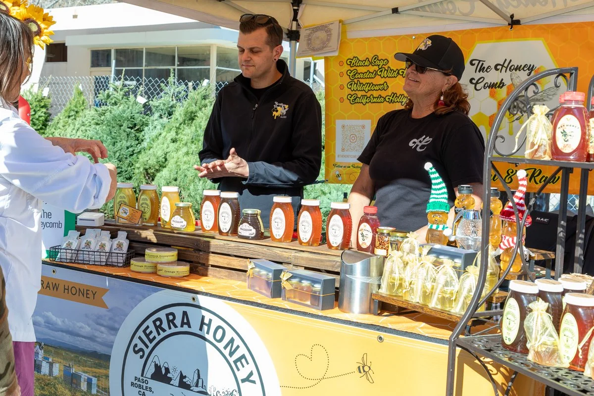 A honey vendor booth at an outdoor market displays jars of honey in front of two smiling women, one with a black cap and sunglasses and the other with a yellow bow headband, while a customer in a white jacket is engaging with them.