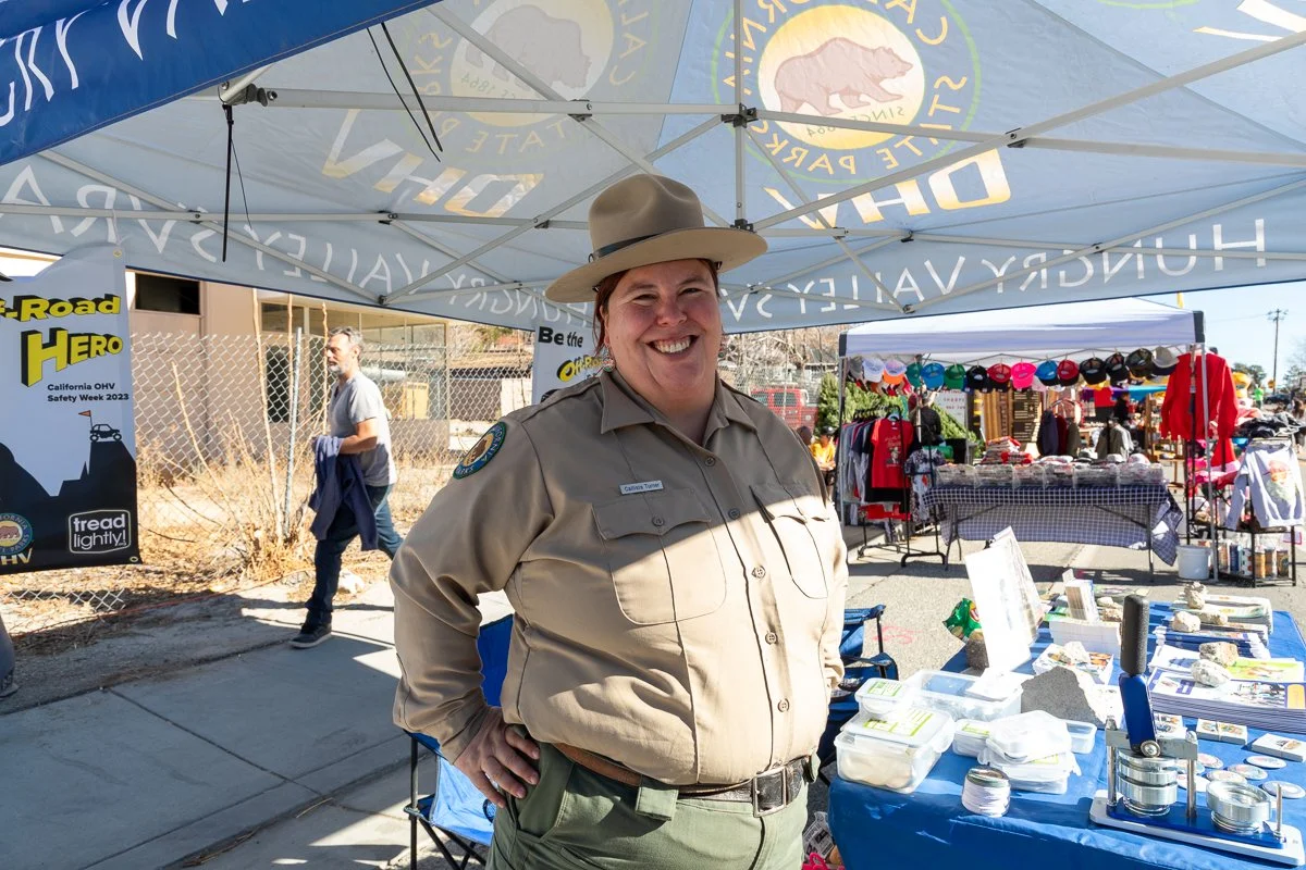 A park ranger in uniform smiling at a safety event booth with outdoor market stalls and a man walking in the background.