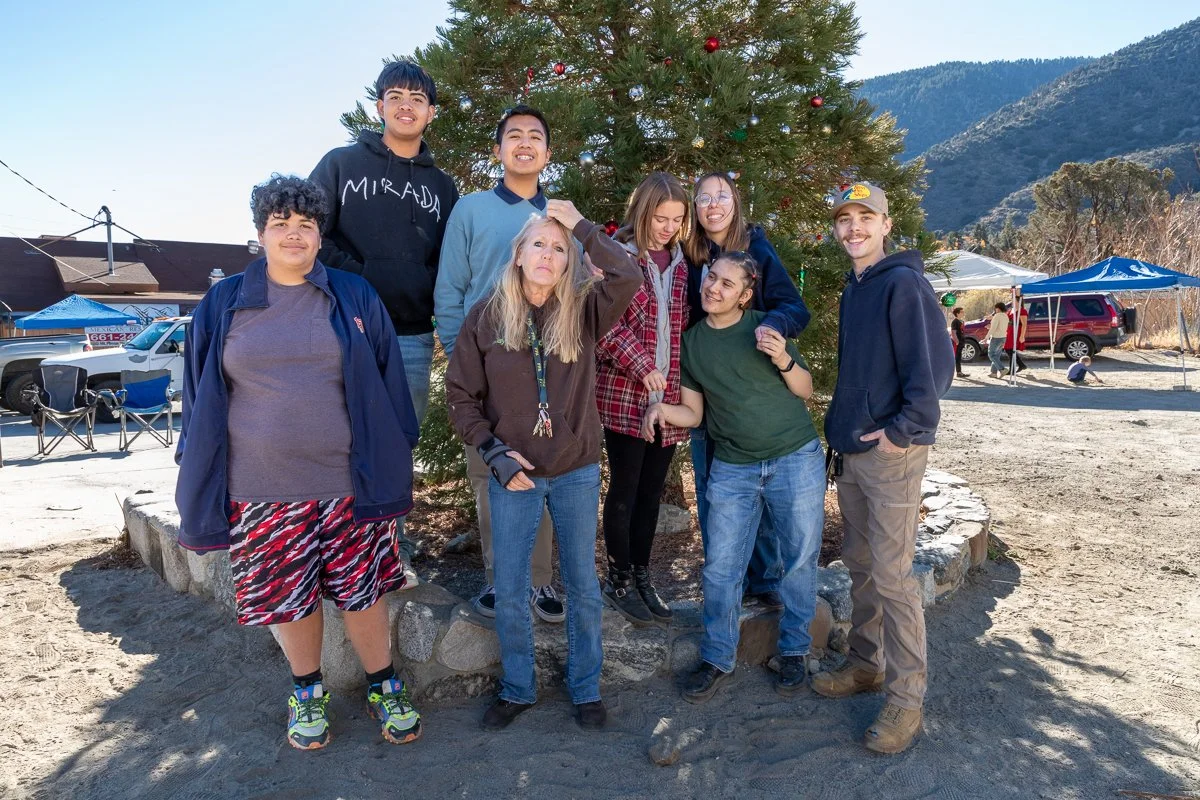 A group of nine people, including children and teenagers, gathered around a decorated Christmas tree outdoors. They are smiling and standing on a circular stone border with mountains and tents in the background.