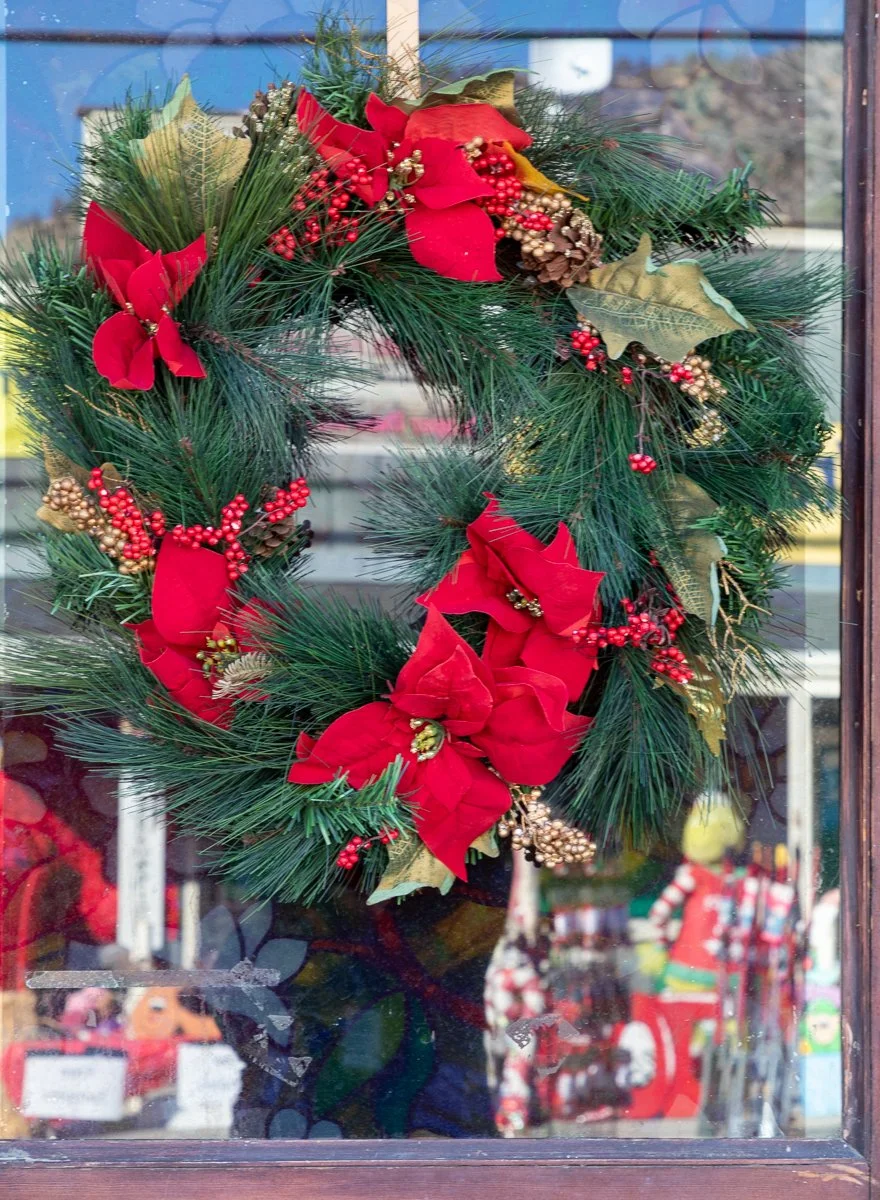 A Christmas wreath decorated with red poinsettia flowers, red berries, pinecones, and green foliage hanging on a glass door.