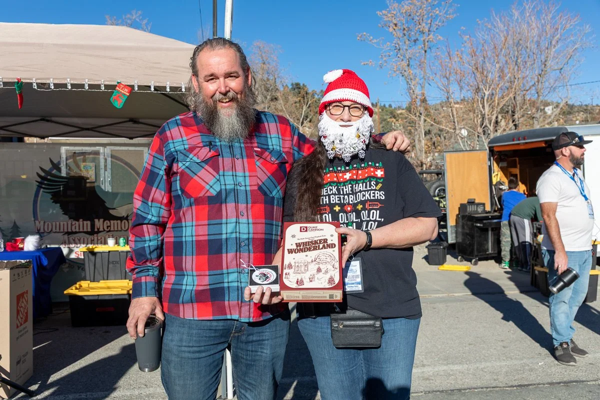 Two people standing outdoors at a festival, one with a gray beard wearing a red and blue plaid shirt, and the other wearing a Santa hat, glasses, a fake white beard, and a black t-shirt, holding a box labeled 'Whisker Wonderland.'