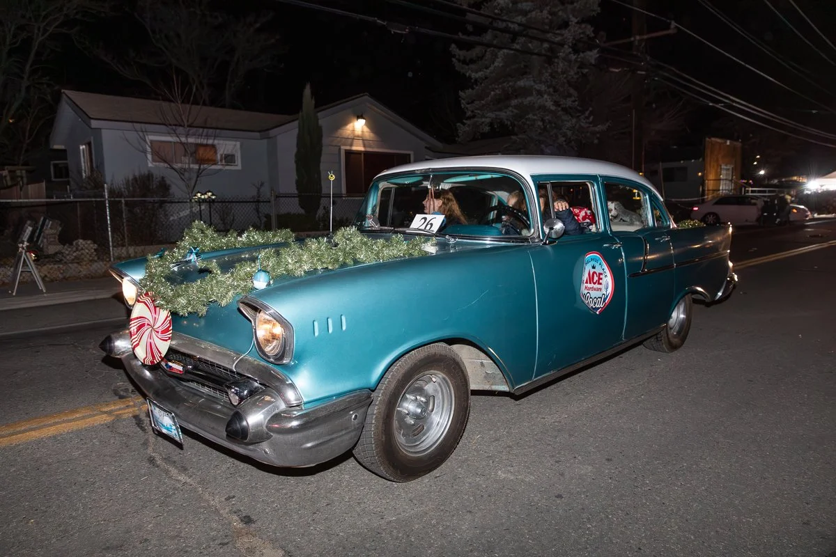 A vintage blue car decorated with Christmas garland and candy cane ornament parked on a street at night.