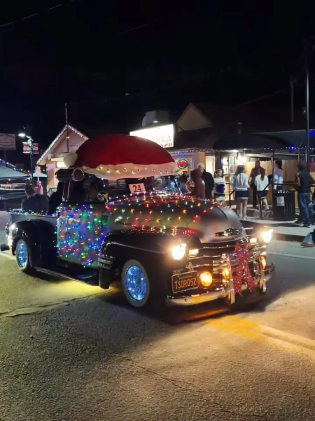 A vintage black car decorated with Christmas lights and ornaments, parked on the street at night during a celebration or parade, with festive decorations and people in the background.