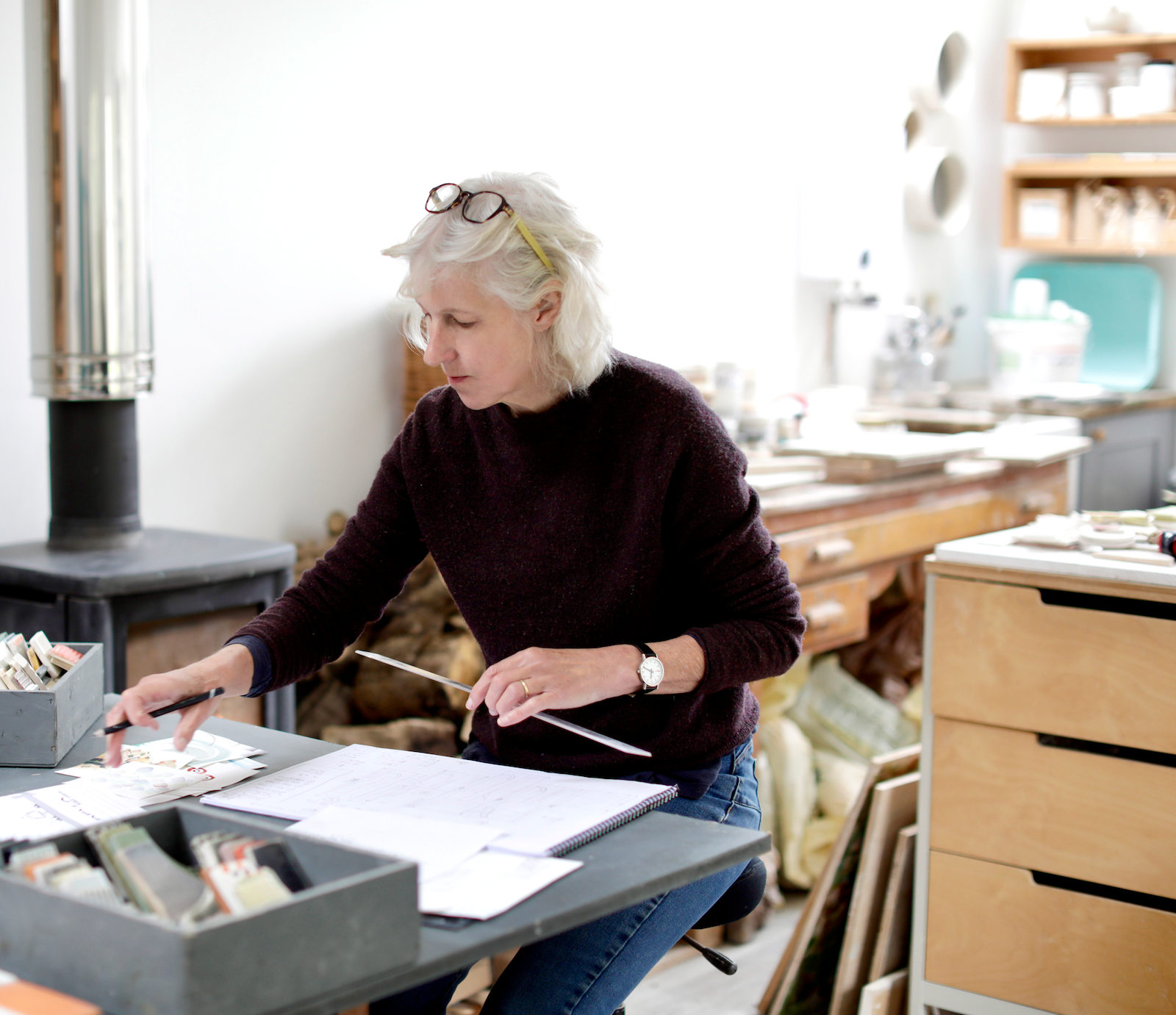 Caroline Egleston is working in an art studio, looking at her supplies and sketches on a table with various art materials and tools around her.