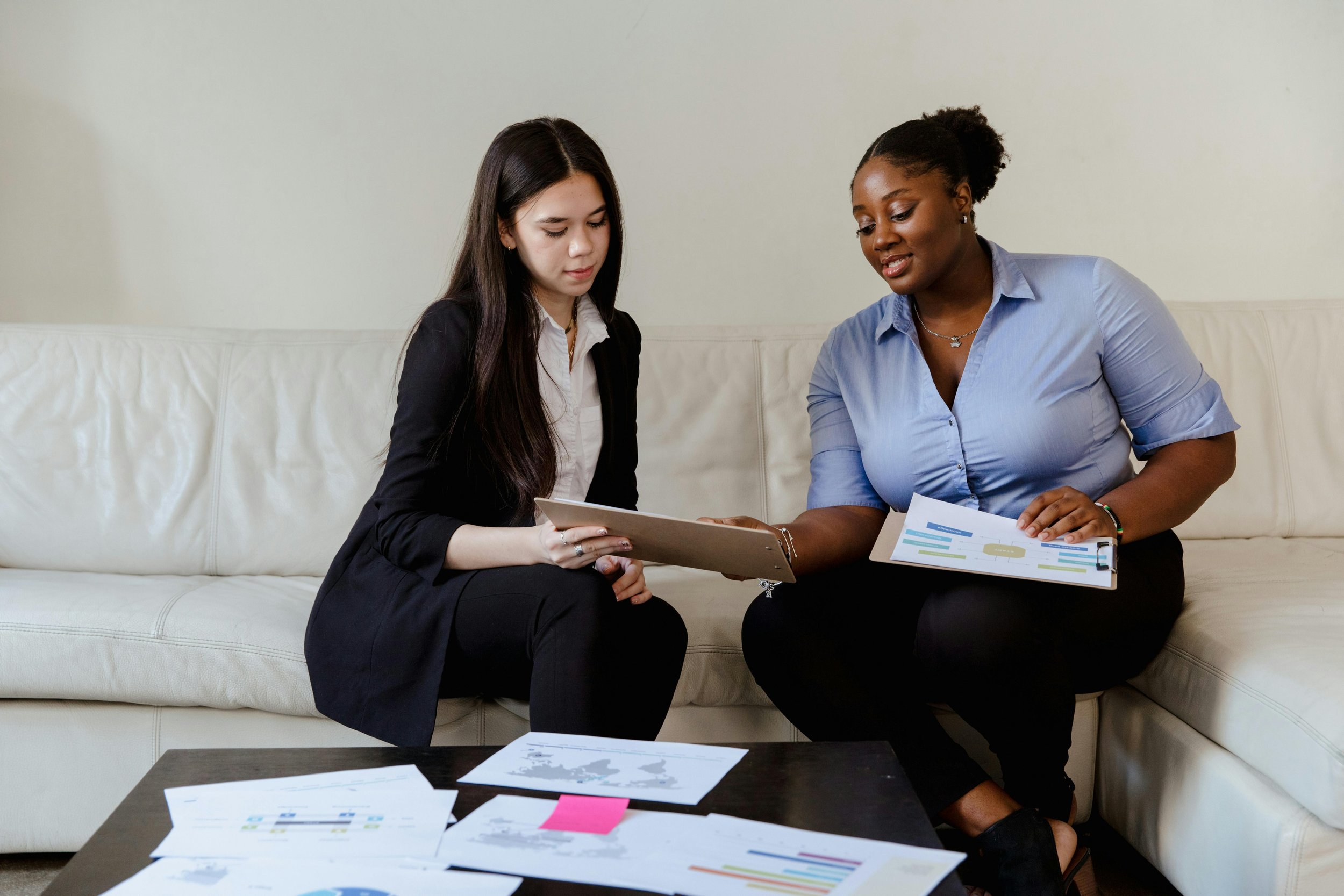 Two women sitting on a white couch discussing documents with charts and graphs, one holding a clipboard and the other holding a printed sheet.
