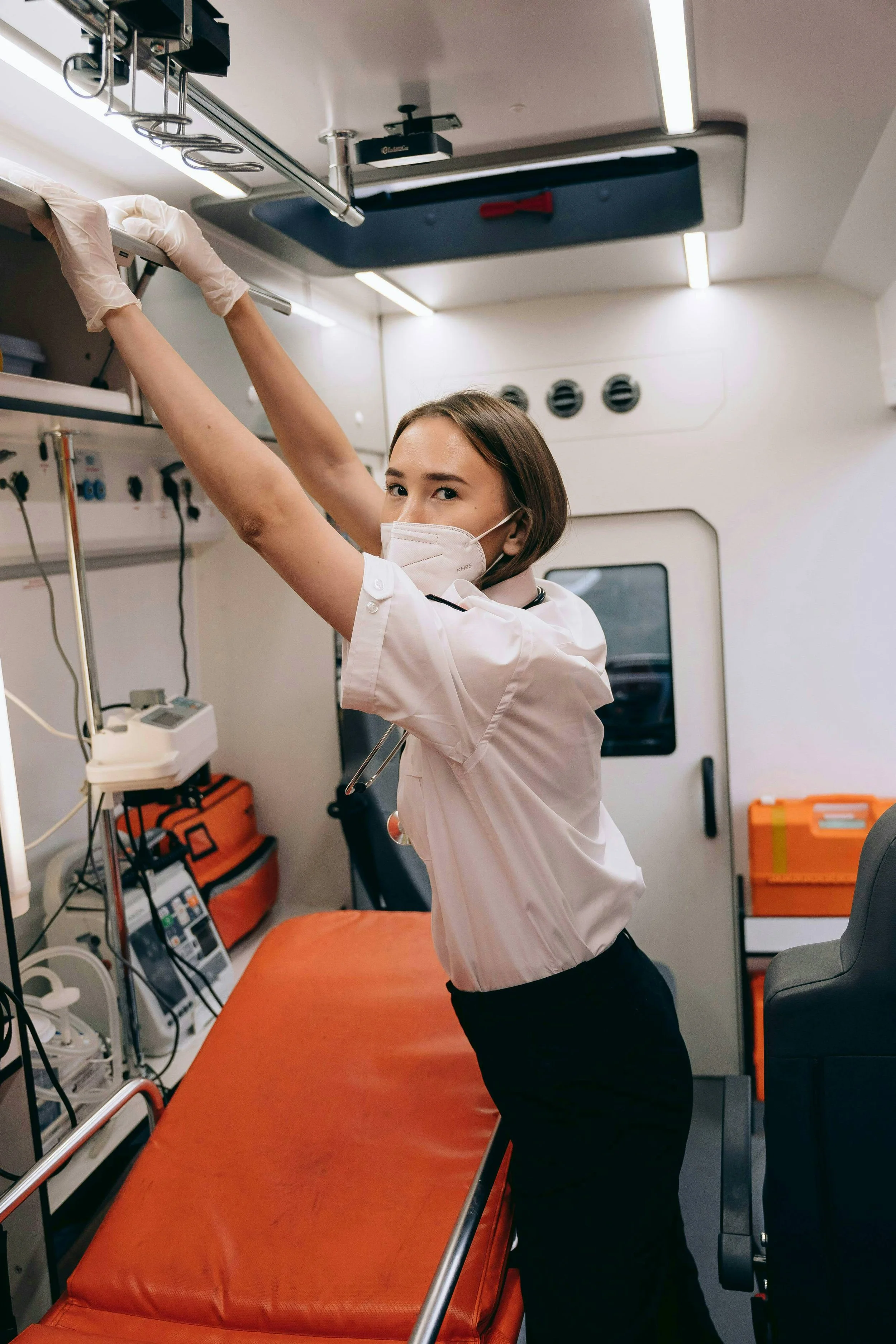 A female medical professional wearing a white uniform, face mask, and gloves, reaching for equipment in a medical emergency vehicle.