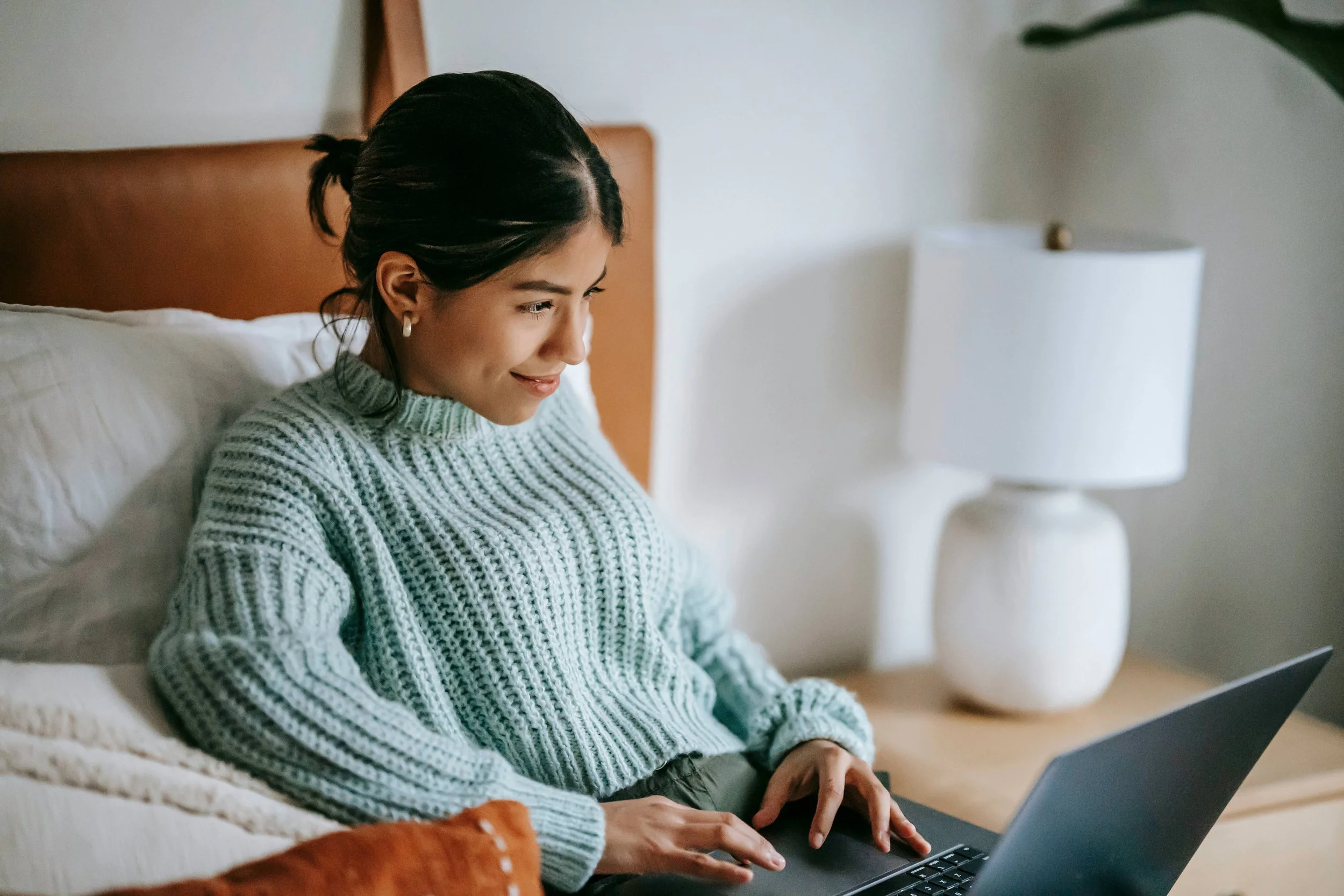 A woman with dark hair tied back, wearing a light green knit sweater, smiling and using a laptop on a beige bed in a cozy, well-lit bedroom.